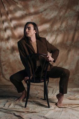 Barefoot queer person in blazer sitting on chair on abstract brown background