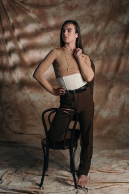 Trendy nonbinary person in corset holding jacket near chair on abstract brown background