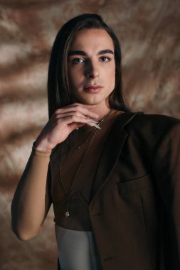 Portrait of stylish nonbinary person touching chin and looking at camera on abstract brown background