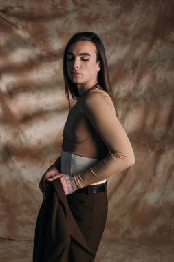 Young nonbinary person holding jacket on abstract brown background