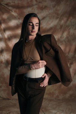Stylish brunette queer person touching jacket on abstract brown background