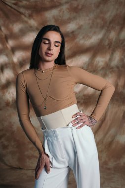 Stylish brunette nonbinary person posing while closing eyes on abstract brown background 