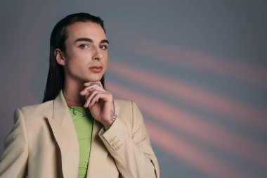 Brunette queer model in jacket looking at camera while touching chin on abstract background 