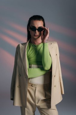 Trendy nonbinary person in sunglasses and suit standing on abstract background 
