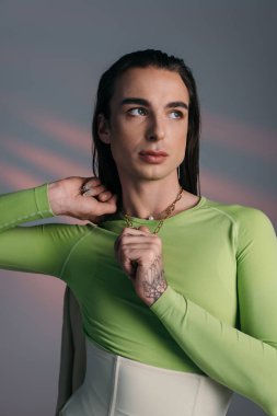 Trendy nonbinary model touching necklace and looking away on abstract background 