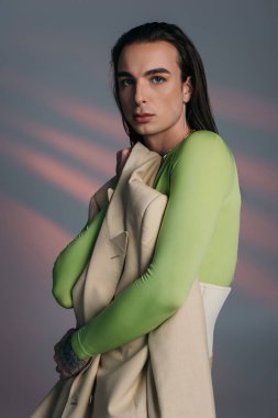 Brunette queer model holding jacket and looking at camera on abstract background 