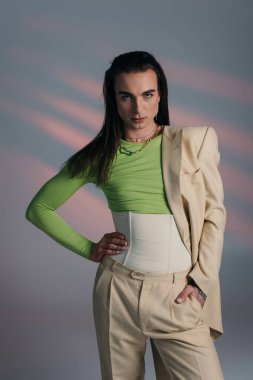 Young tattooed queer model in suit and corset posing on abstract background 