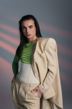 Stylish queer model in suit and corset standing on abstract background 