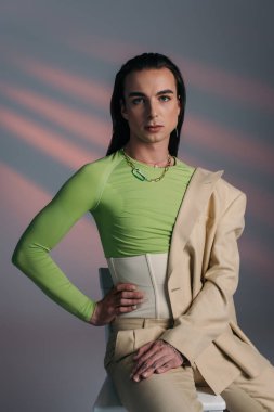 Trendy young queer model in suit and corset posing on chair on abstract background 