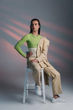 Stylish nonbinary model posing while sitting on chair on abstract background 