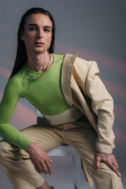 Portrait of trendy queer model in beige suit sitting on chair on abstract background 