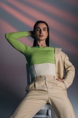 Stylish queer model in beige suit and corset sitting on chair on abstract background 