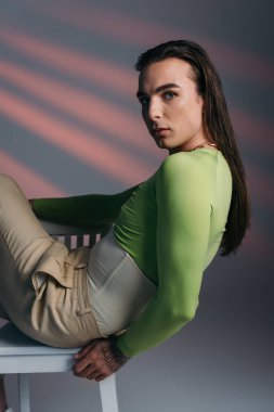 Trendy brunette nonbinary model sitting on chair and looking at camera on abstract background 