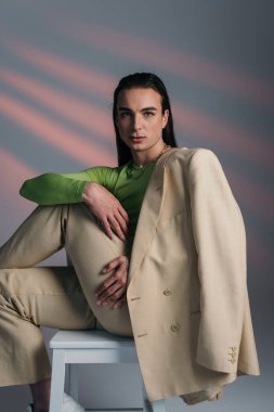 Fashionable queer model in suit posing on chair and looking at camera on abstract background 