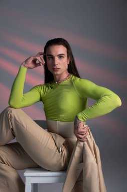 Young brunette queer person holding jacket while sitting on chair on abstract background 