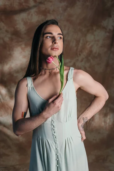 Brunette nonbinary person in sundress holding tulip on abstract brown background