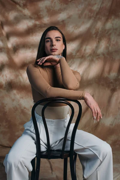 Young queer person in corset and white pants sitting on chair on abstract brown background