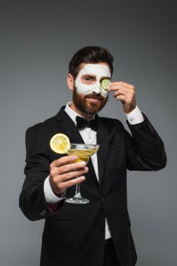 happy man in suit with clay mask on face holding cocktail isolated on grey 