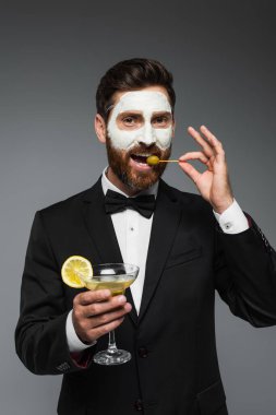 bearded man in suit with clay mask on face holding cocktail and eating olive isolated on grey 