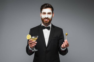 happy man in suit with clay mask on face holding cocktail and toothpick with olive isolated on grey 
