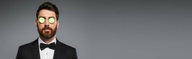 bearded man in tuxedo with fresh sliced cucumber on eyes isolated on grey, banner