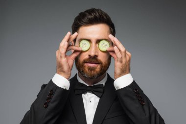 bearded man in tuxedo holding fresh sliced cucumber near eyes isolated on grey