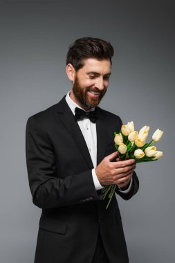 happy bearded man in elegant tuxedo with bow tie holding fresh tulips isolated on grey 