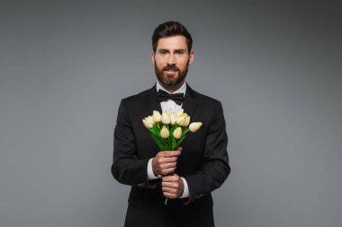 bearded man in elegant suit with bow tie holding bouquet of tulips isolated on grey 