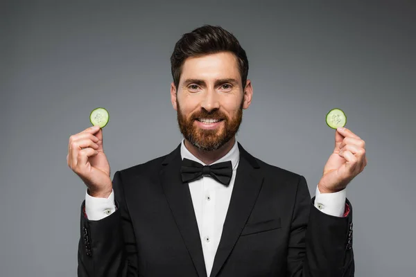 bearded man in tuxedo holding fresh sliced cucumber and smiling isolated on grey