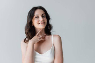 Happy brunette woman looking at camera and touching neck isolated on grey 