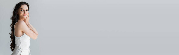 Positive model in white clothes smiling at camera isolated on grey, banner  