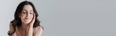 Positive brunette woman with closed eyes touching face isolated on grey, banner 