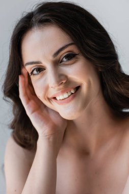 Cheerful brunette woman touching face isolated on grey 
