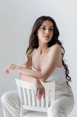 Brunette woman in white clothes sitting on chair isolated on grey 