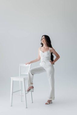 Smiling woman in white clothes posing near chair on grey background