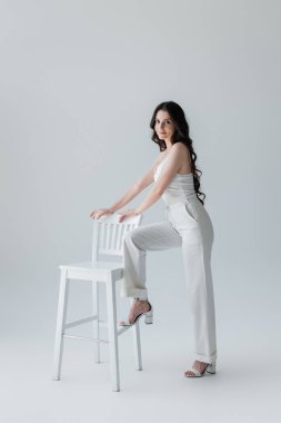 Smiling model in heels and white clothes looking at camera near chair on grey background