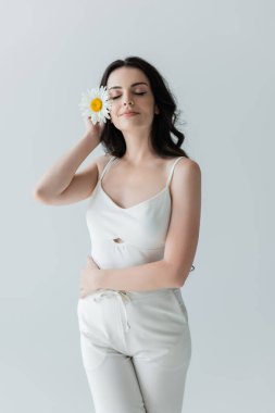 Pleased woman in white clothes holding chamomile isolated on grey 