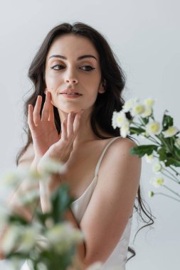 Young woman touching face and looking at blurred flowers isolated on grey 