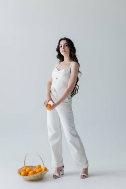 Stylish young woman holding orange near wicker basket on grey background