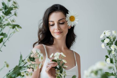 Smiling woman in white top touching flowers isolated on grey 