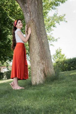 Stylish woman in blouse and skirt standing near tree in park 