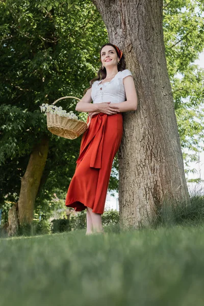 Low angle view of smiling woman in blouse and skirt holding basket with flowers in park