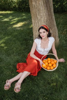 High angle view of trendy woman sitting near oranges in basket and tree in park 
