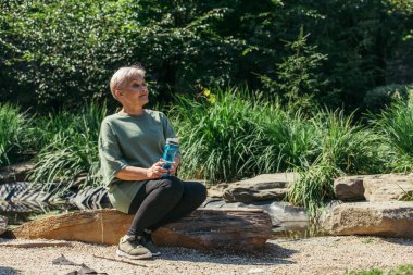 retired woman in sportswear sitting with sports bottle on wooden log