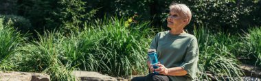 retired woman in sportswear sitting with sports bottle around green plants, banner