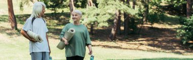 cheerful senior couple in sportswear holding fitness mats and sports bottles in park, banner