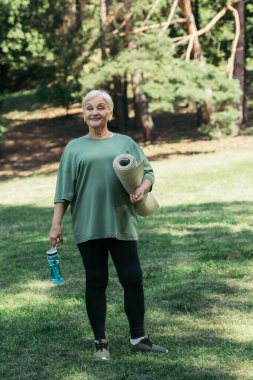 full length of happy senior woman holding sports bottle and fitness mat in park