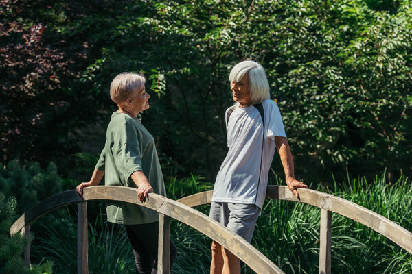 retired senior couple in sportswear looking at each other while standing on bridge 