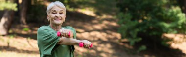 happy senior woman with grey hair smiling and exercising with dumbbells in park, banner