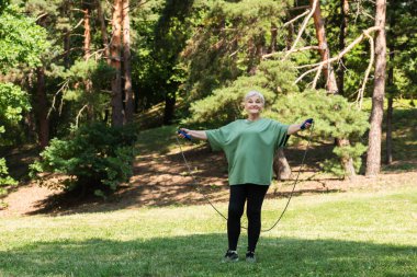 full length of senior woman with grey hair smiling and exercising with jumping rope in park